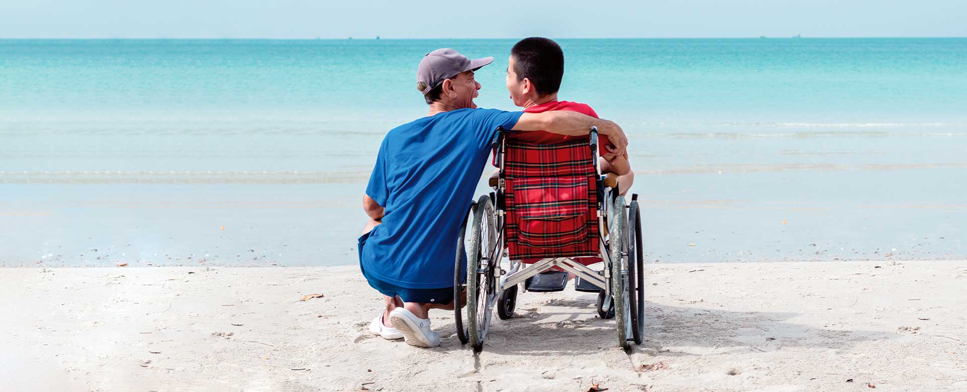 Un uomo in spiaggia abbraccia un ragazzo su sedia a rotelle di fronte al mare 